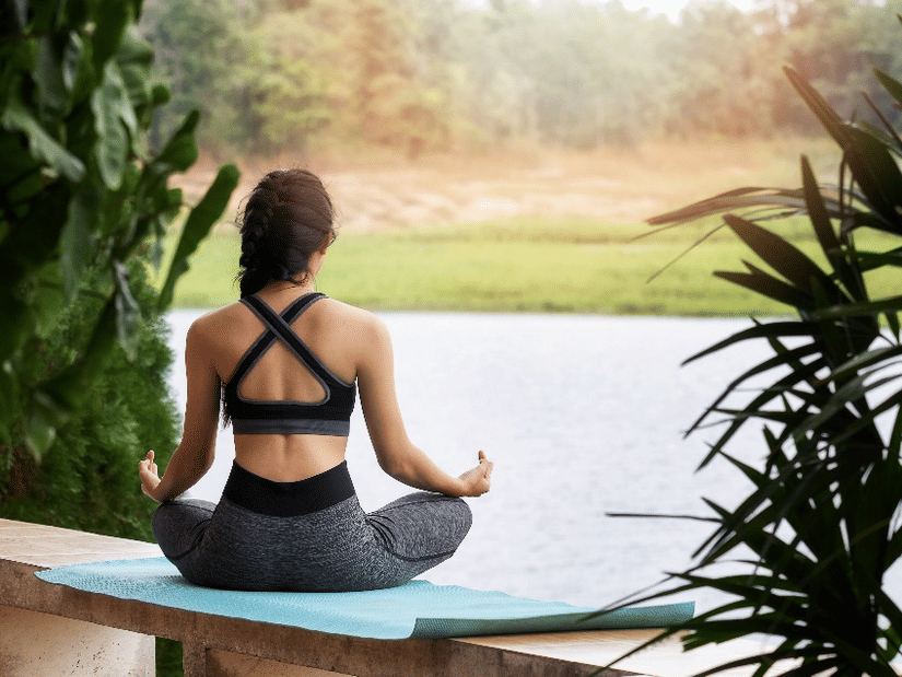 A woman practices seated meditation on a yoga mat by a peaceful lakeside, surrounded by lush greenery and calming natural views