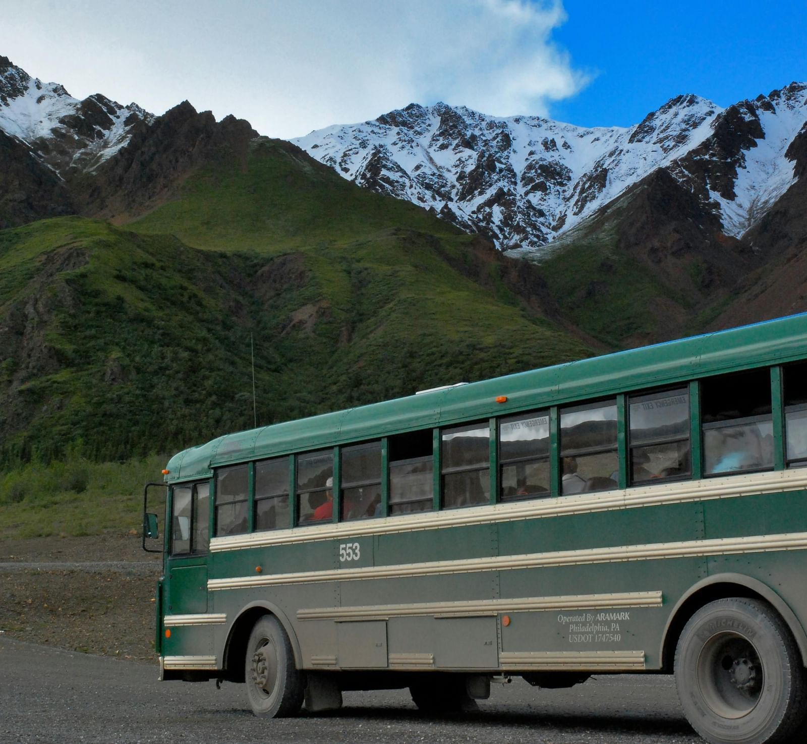 Green bus travelling through a road with green mountains in the background and snow on peak of the mountains.
