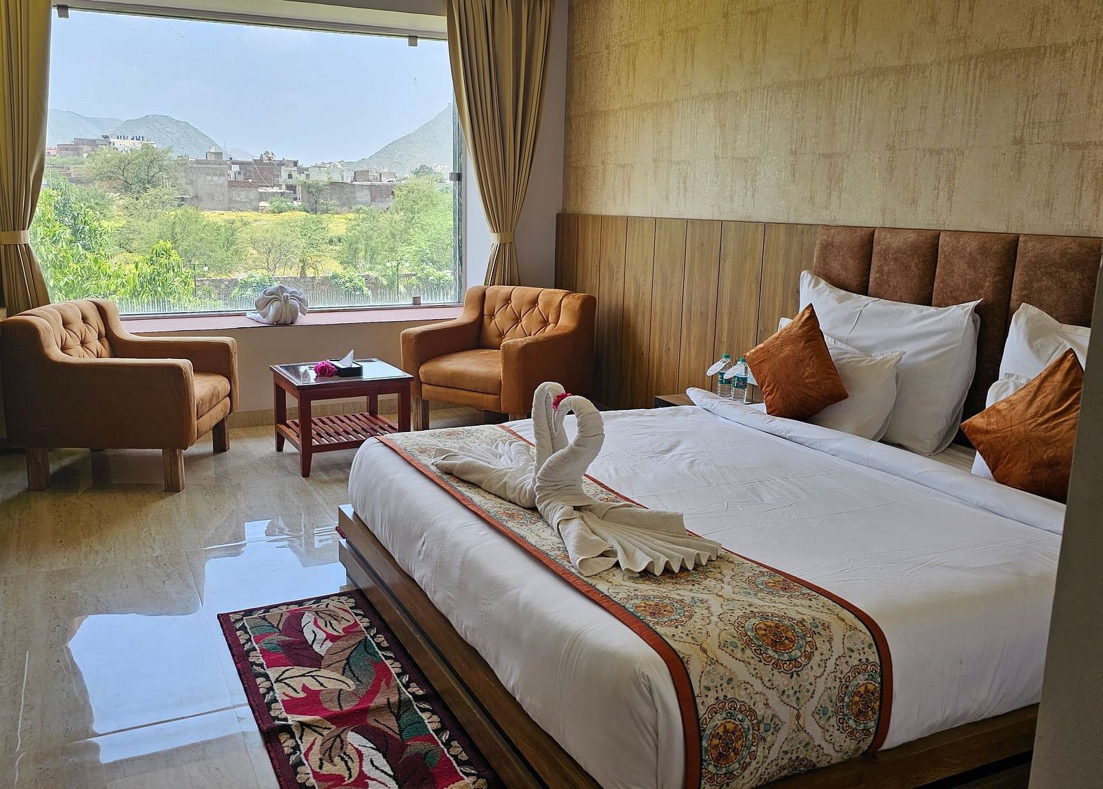 A bright and spacious resort room at Bamboo Saa Mulberry Resort, Pushkar, featuring a large window with a view of the outdoors including a made bed with a patterned runner, two armchairs, and a small table.