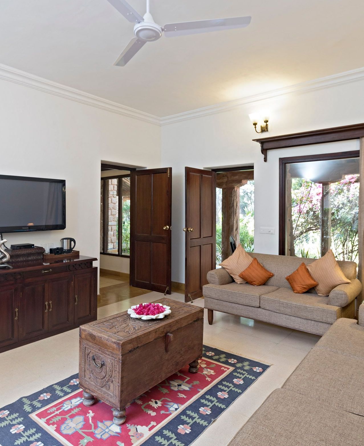 A living room with two sofas, wooden center table on patterned rug, TV on cabinet, floor vase, table lamp, cushions, ceiling fan, and doors opening outside - MANVÂR Shergarh The Desert Resort