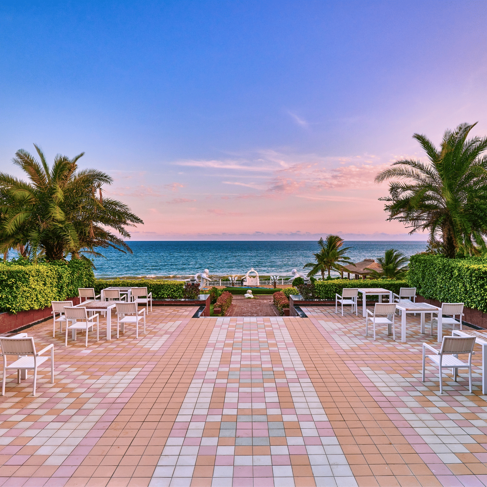 Open tiled terrace with white tables and chairs, clipped hedges and palm trees overlooking the sea at dawn at MAYFAIR Palm Beach Resort, Gopalpur-on-Sea.