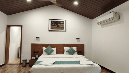 A hotel room with king bed, teal accent pillows, wooden headboard, vaulted ceiling, wall AC, and wildlife artwork above the bed.