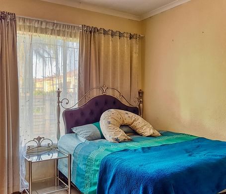 A bedroom featuring a double bed with blue sheets spread neatly over it with beige curtains covering the window behind it.