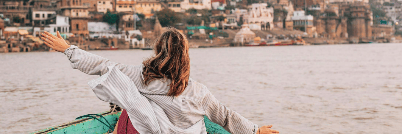 A woman sitting on a wooden boat with arms outstretched, facing a riverfront city with buildings, temple structures, water in the foreground.