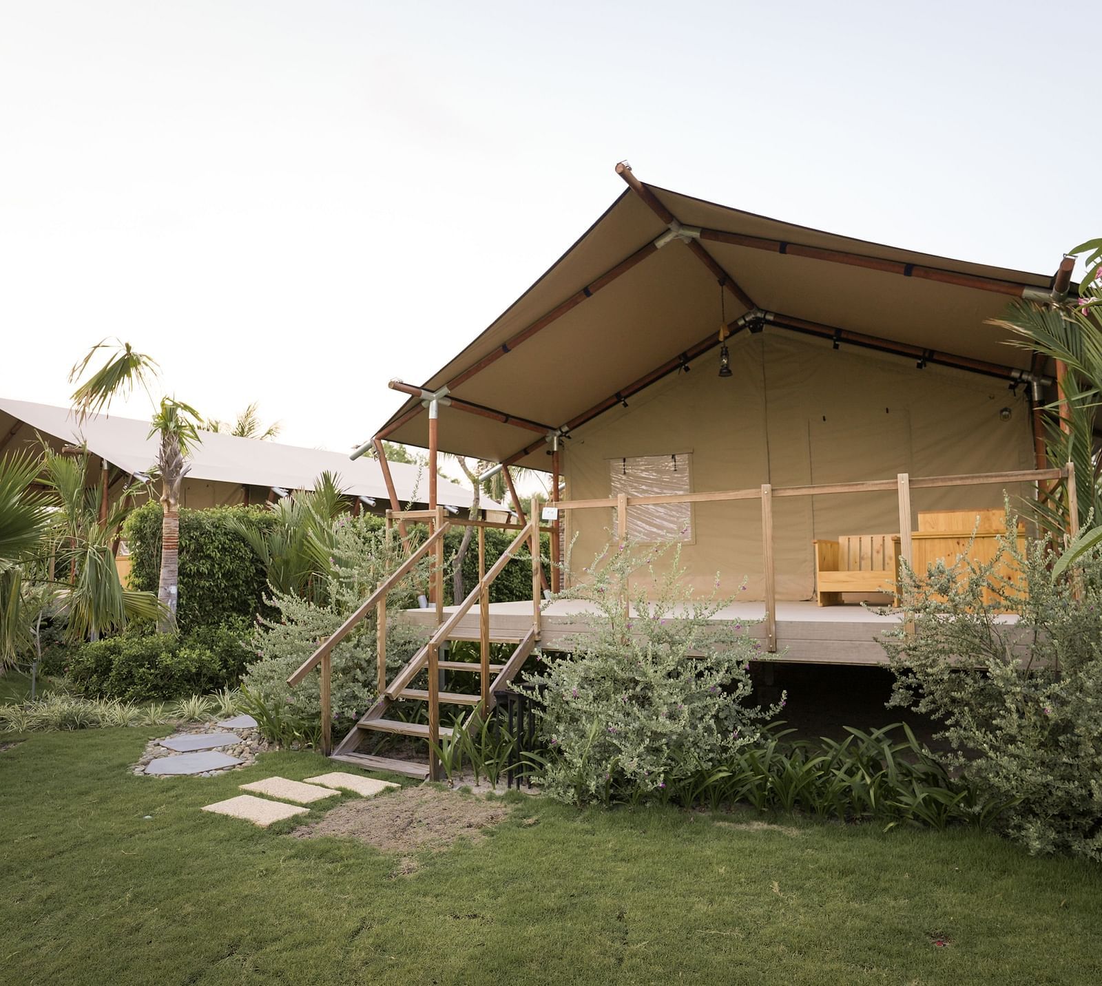 A front-facing view of the Peaceful Tent at WoCAL Retreat Ke Ga, showcasing a wooden staircase that leads to a furnished deck, surrounded by manicured garden beds and tropical vegetation.
