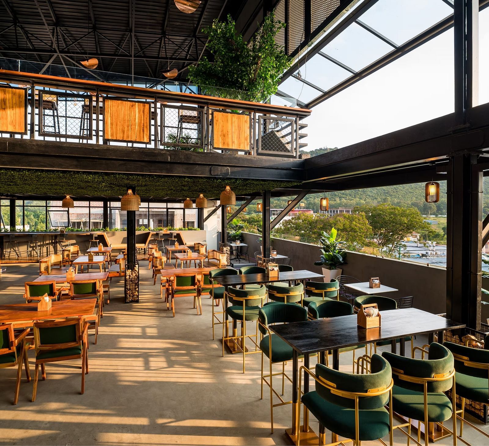 A spacious rooftop restaurant with elegant wooden furnishings, green velvet chairs, and panoramic views of the hills, bathed in warm natural light at TGI Livancia Hillside, JC Nagar, Mysore.