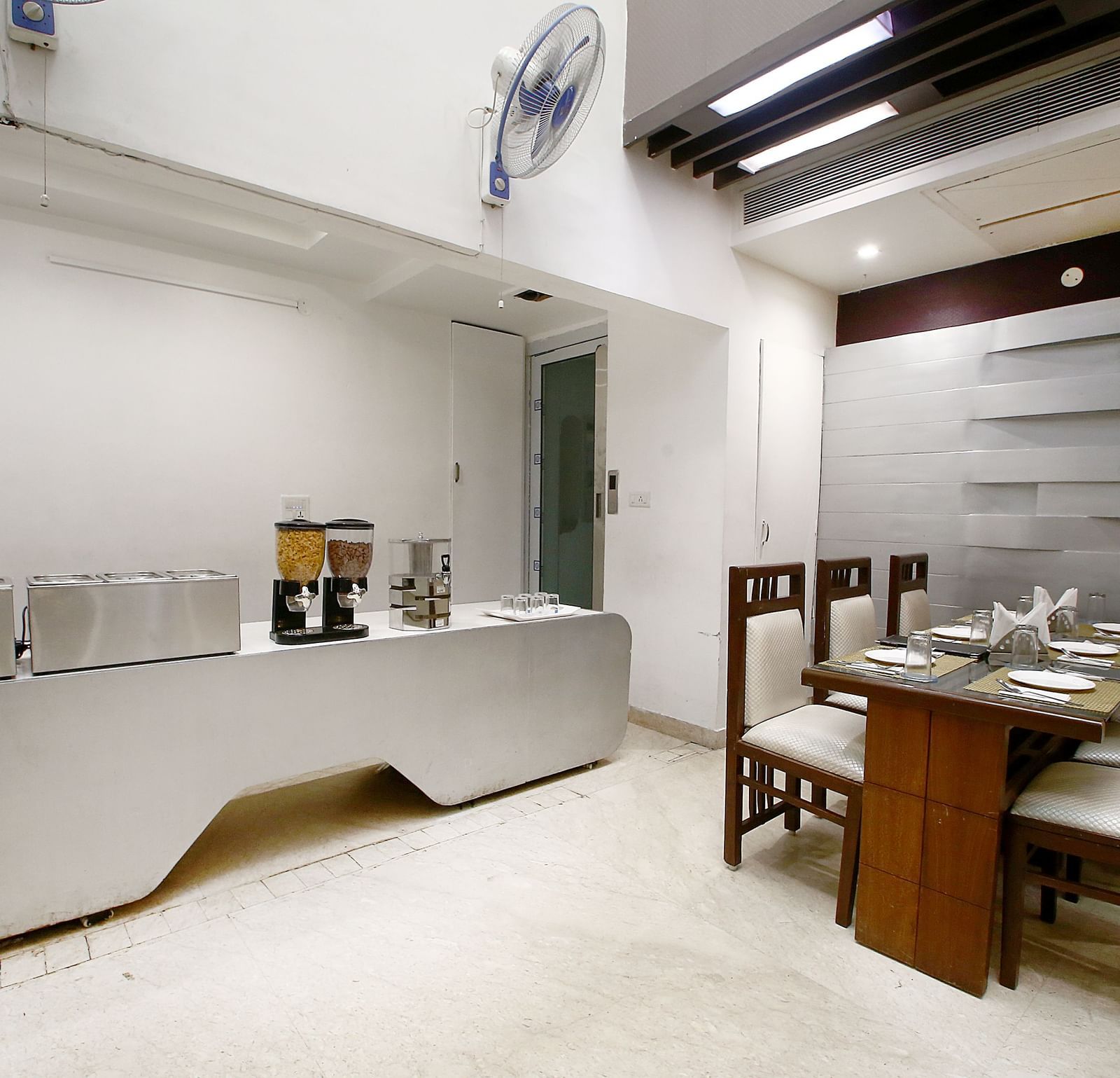 Dining space with a table and four chairs around it in a corner, a table with white plates and a desk with utensils along with two fans in a modern interior setting - Hotel Emerald, Chandigarh