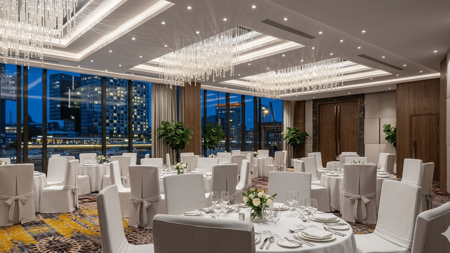 A bright banquet hall at KPG Sarovar Portico, Raj Nagar Extension, Ghaziabad, set with many round tables and white-covered chairs, featuring large windows and crystal lighting.