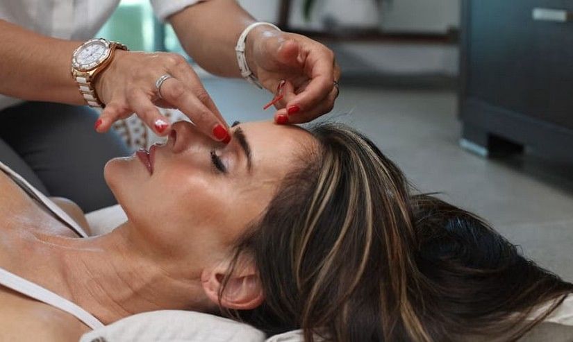 A woman with brown hair lies down, receiving a facial treatment with hands gently touching her forehead.