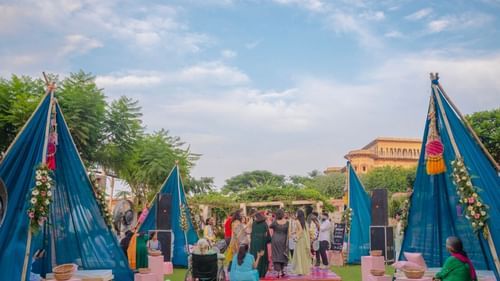 Blue tents and seating are arranged on a green lawn for an event at Tijara Fort-Palace - 19th Century, Alwar under a partly cloudy sky.