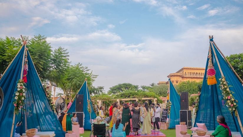 Blue tents and seating are arranged on a green lawn for an event at Tijara Fort-Palace - 19th Century, Alwar under a partly cloudy sky.