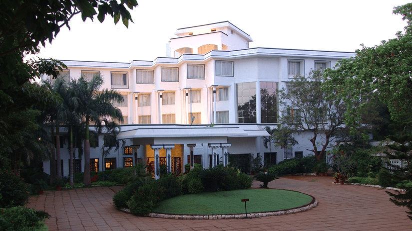 Facade view of Sangam Hotel, Thanjavur with a pathway, a manicured garden and trees in view - Thanjavur Hotel Contact