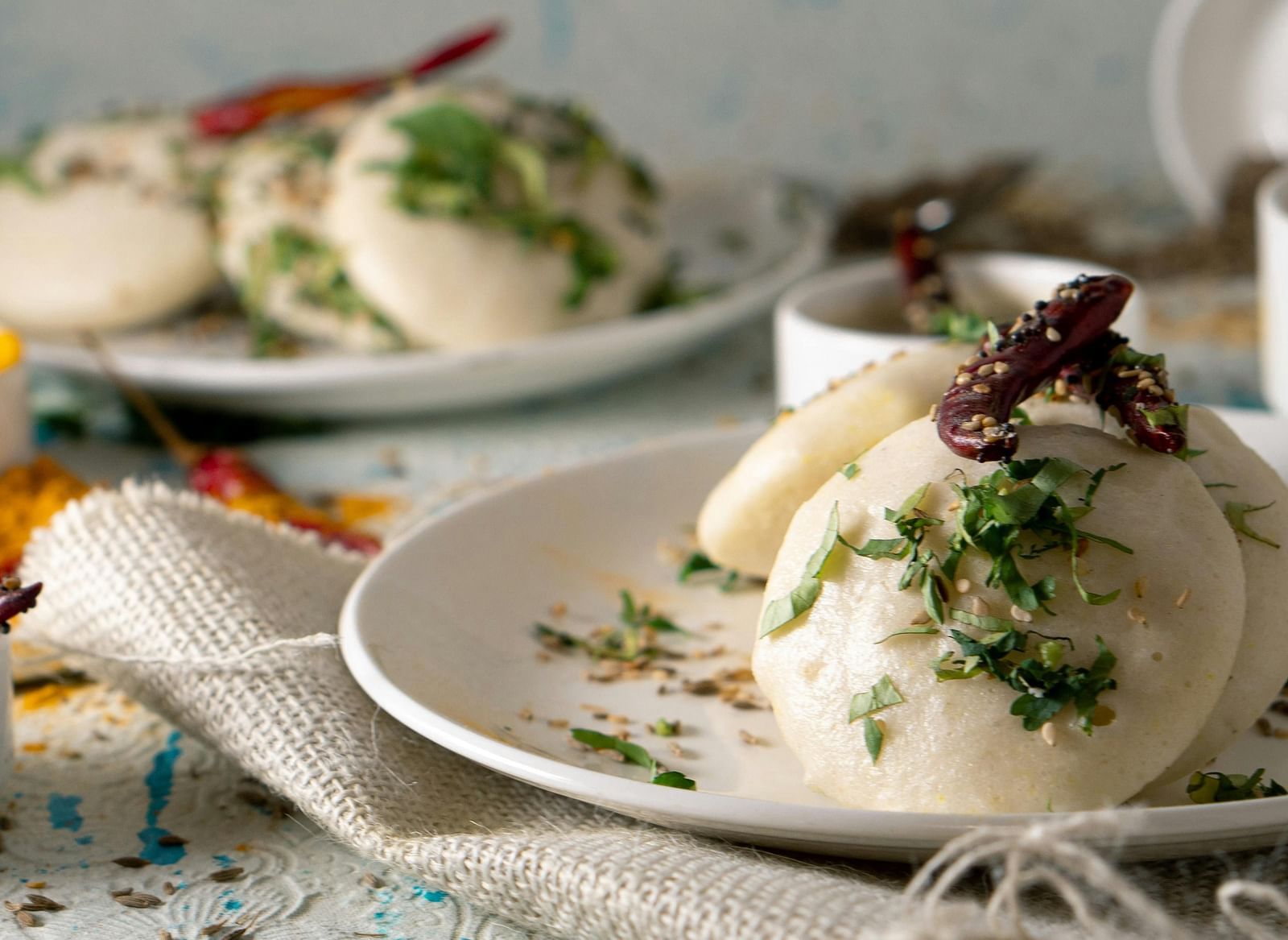 Steamed idlis garnished with herbs and chilli flakes are presented on a plate, with more idlis and dipping sauces in the background.