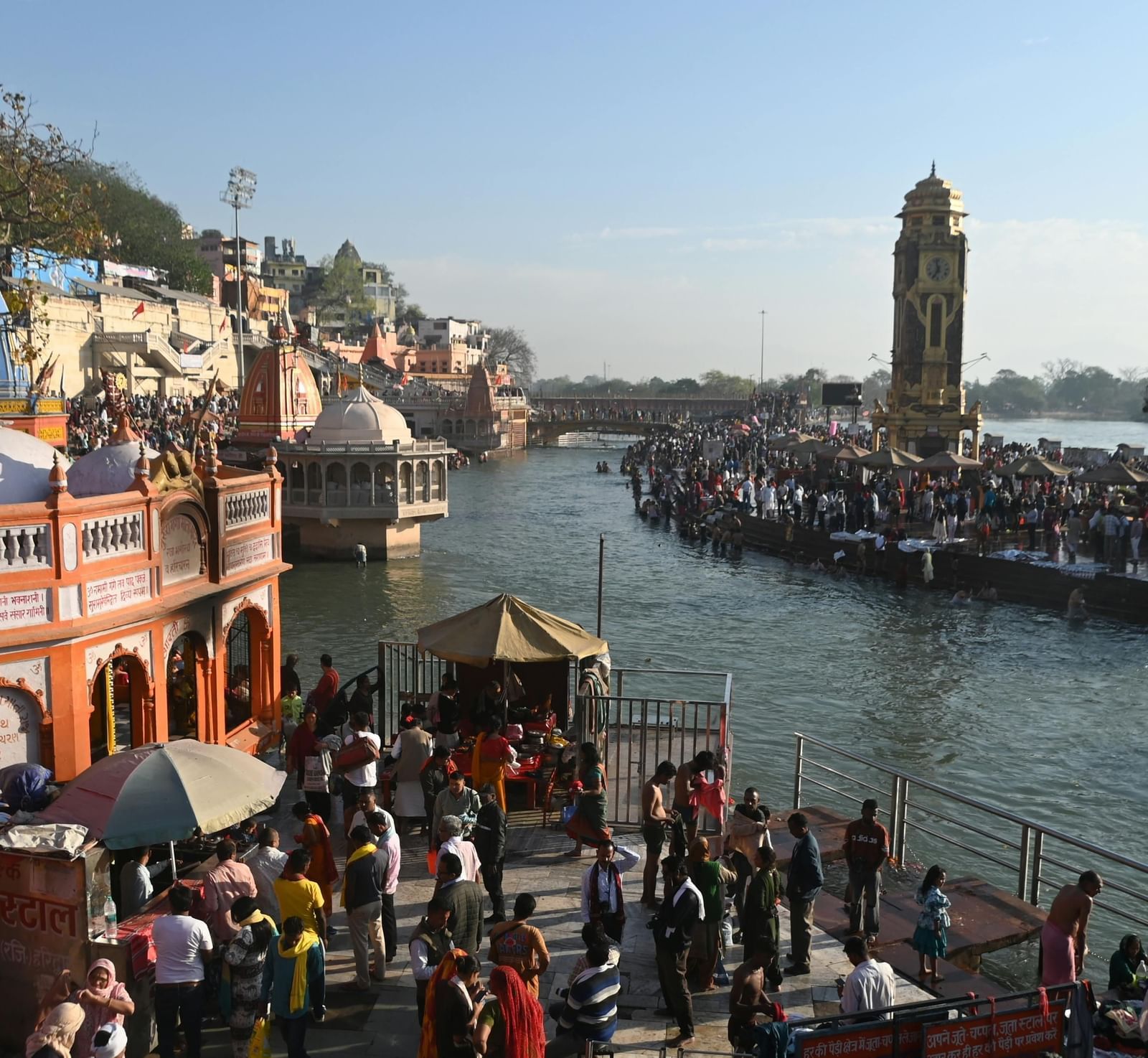 An expansive view of the crowded Har Ki Pauri ghat in Haridwar, with the River Ganges flowing through the center, flanked by temples, the tall Ghanta Ghar clock tower, and numerous pilgrims.