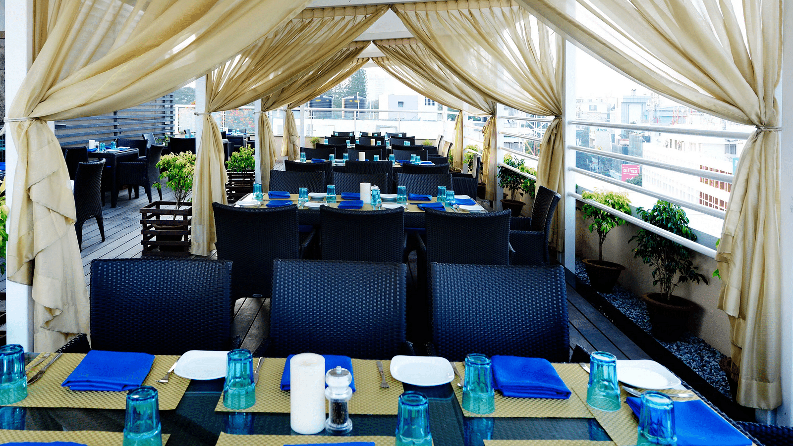 Open-air dining space with elegant golden drapes, black chairs, and neatly set tables with blue accents at Skylit Rooftop Restaurant, The Cunningham Hotel