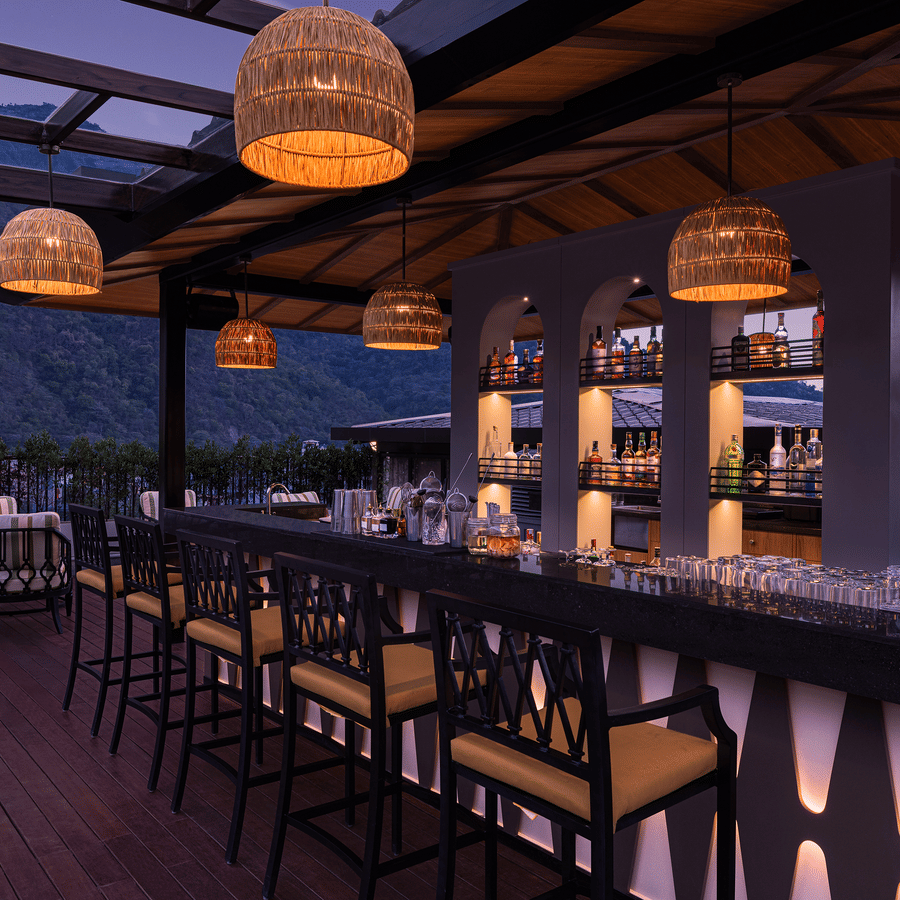 Contemporary bar at The Terrace, Aloha on the Ganges, Rishikesh, featuring high bar stools, illuminated shelving, and panoramic mountain views through glass walls.