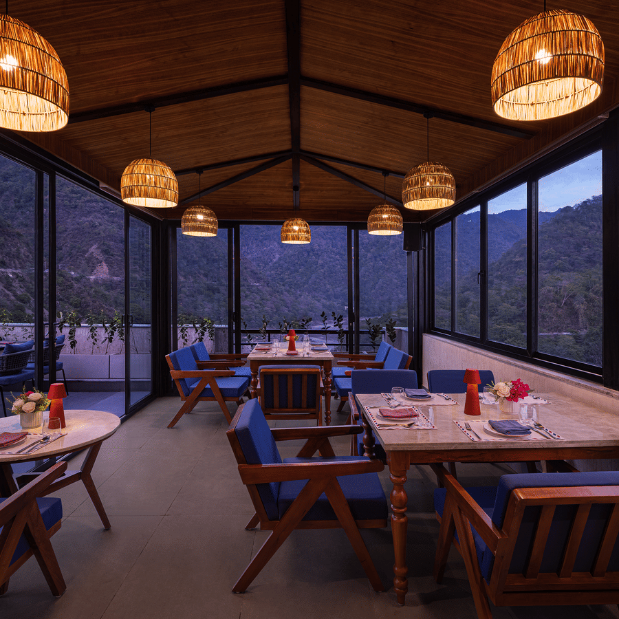 Indoor dining area at The Terrace, Aloha on the Ganges, Rishikesh, featuring wooden tables, pendant lighting, and floor to ceiling glass windows overlooking the hills.