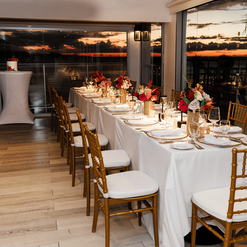 A long dining table elegantly set with white linens, gold chairs, and floral centrepieces inside a glass-walled venue, overlooking a scenic sunset view.