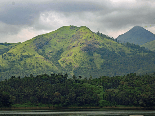 A view from afar of a forest cover with a large green hill having trees behind it, and other peaks in the background as seen during a cloudy day.