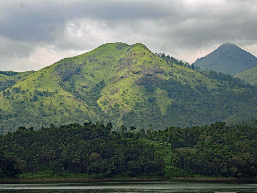 A view from afar of a forest cover with a large green hill having trees behind it, and other peaks in the background as seen during a cloudy day.