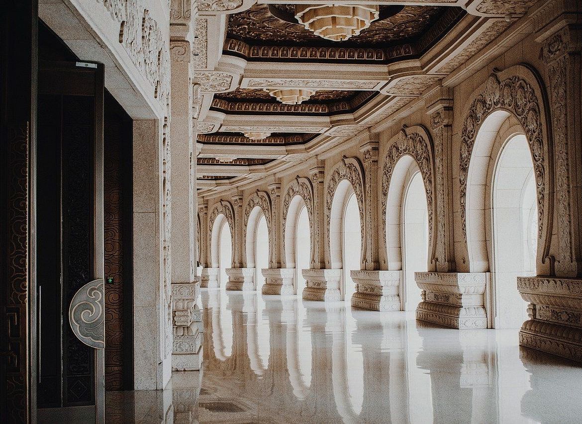 A luxurious hallway with arched windows, columns, and a polished marble floor, evoking a sense of grandeur and opulence.