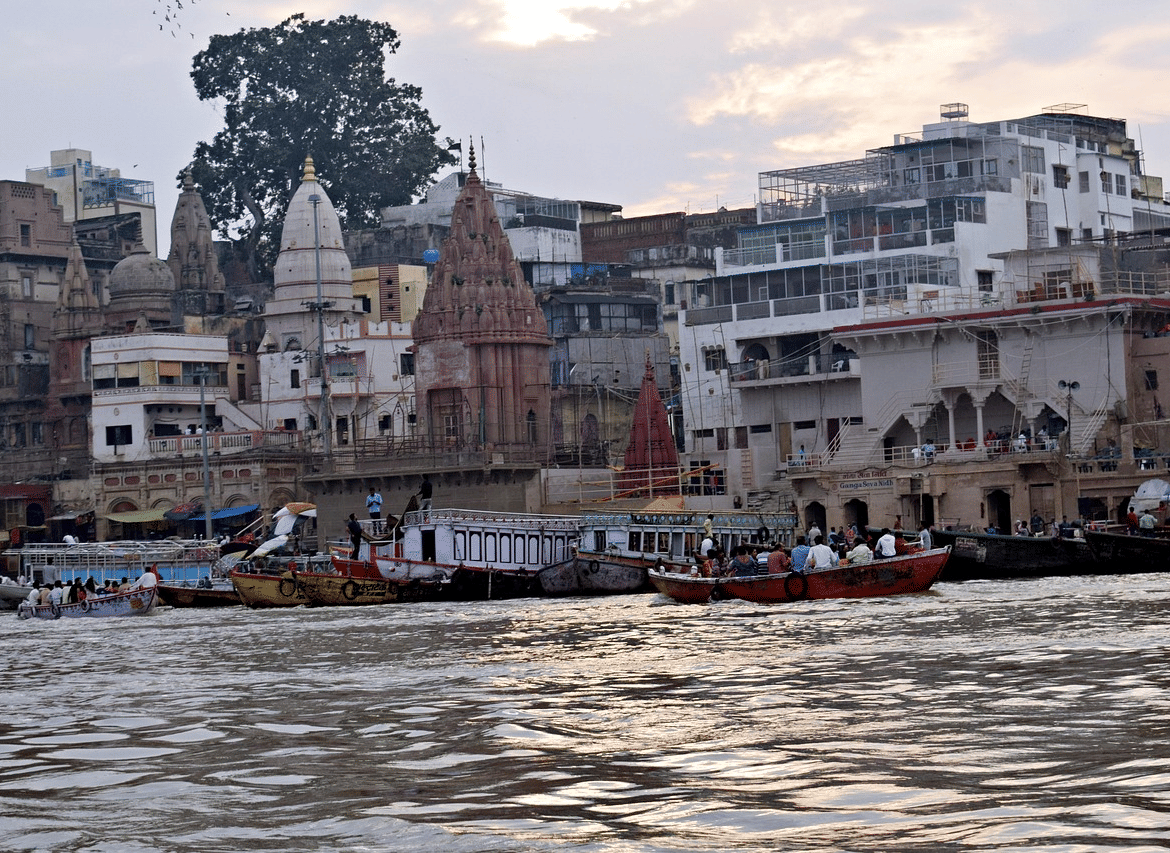 A ghat near the Ganges River showing boats moving on the water, with temples and buildings lining the bank, under an overcast sky.