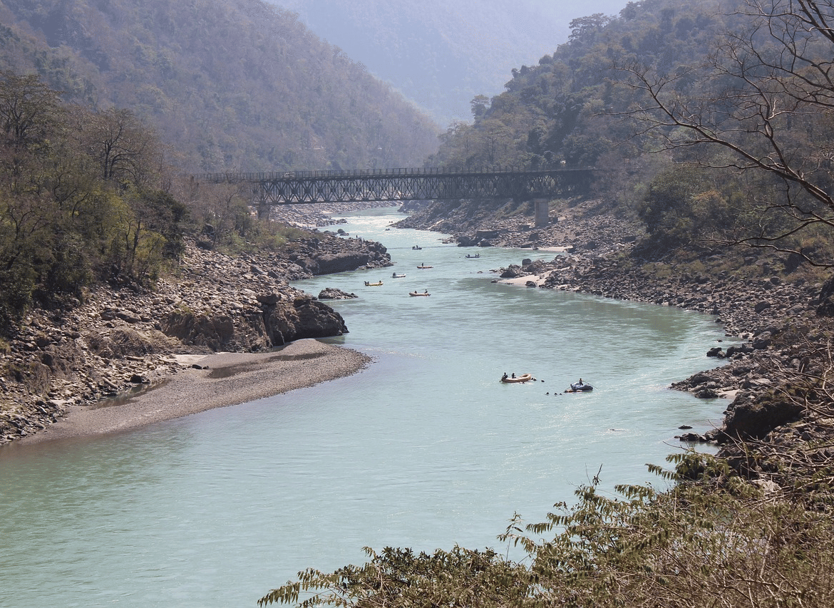 A wide, winding river flowing through a rocky gorge with a bridge visible in the distance, surrounded by green hills