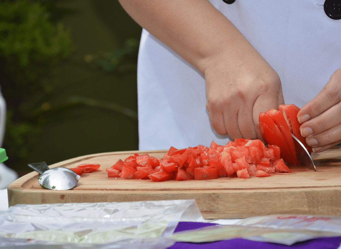 A person wearing a chef coat chopping tomatoes on a wooden cutting board with a spoon and packets nearby.