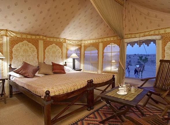 An image of the inside of a tent with a twin bed, a wooden chair and table, bedside tables and tables - MANVAR Resort &Desert Camp
