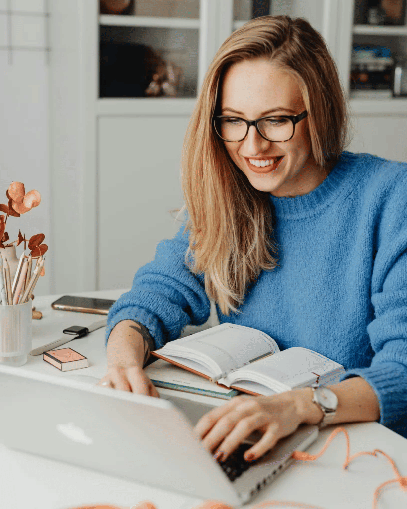 A smiling woman with glasses and a blue sweater working on her laptop at a white desk, with an open notebook and desk accessories nearby.