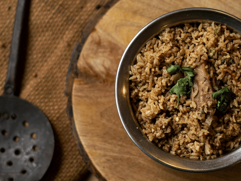 Bowl of Chettinad-style rice placed on a wooden surface, garnished with curry leaves, accompanied by a traditional metal utensil