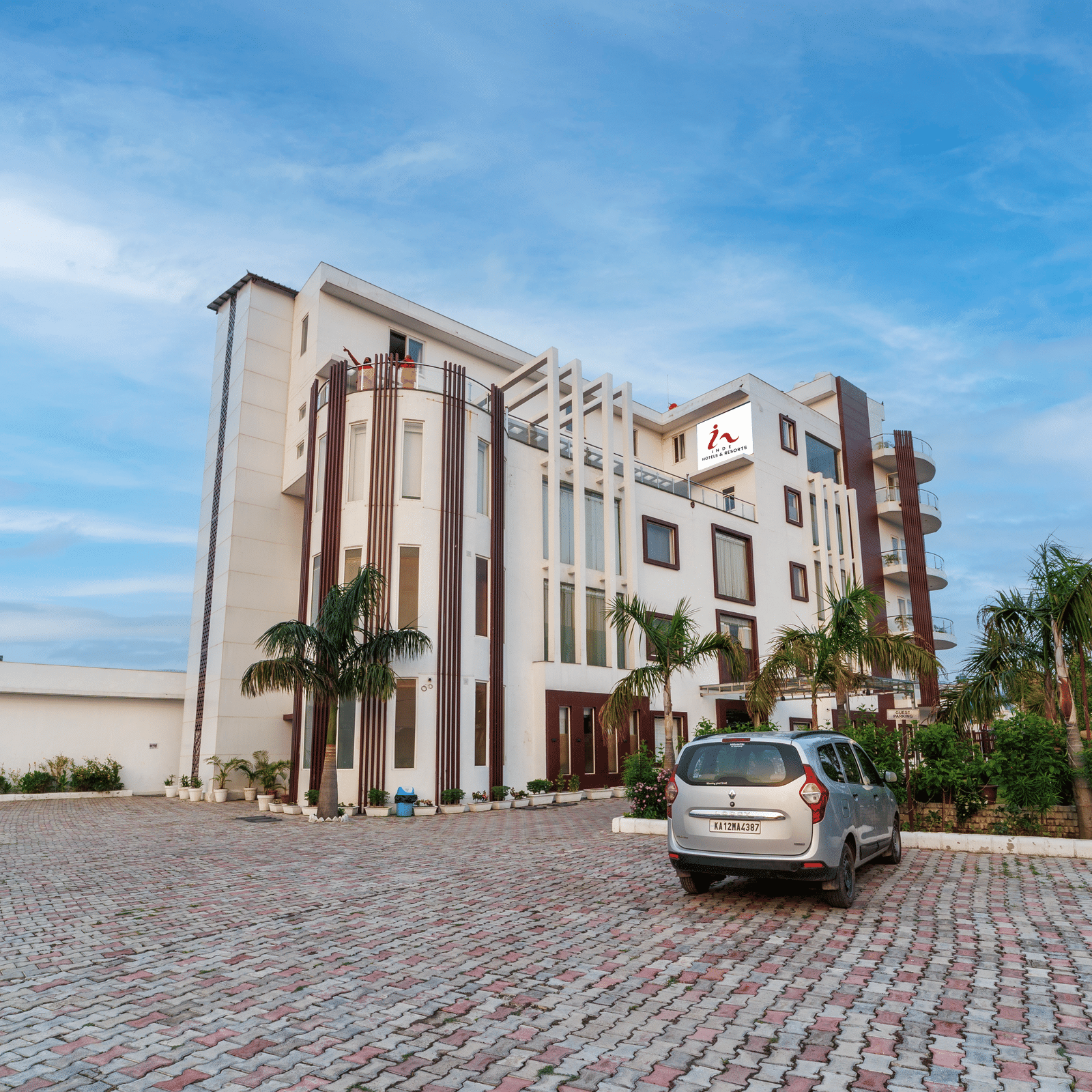 Exterior view of Inde Reva Resorts Rishhikesh with paved driveway, parked car and palm trees.