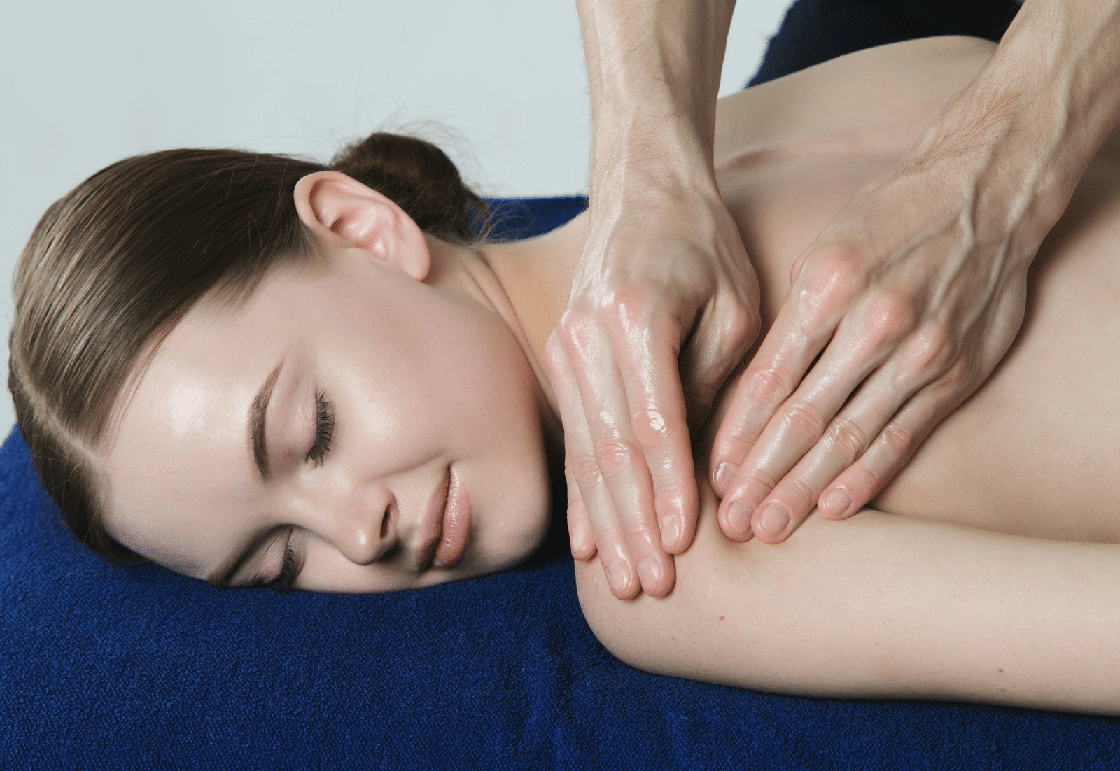 A massage therapist uses oiled hands to massage the shoulder and upper back of a woman lying on a massage table.