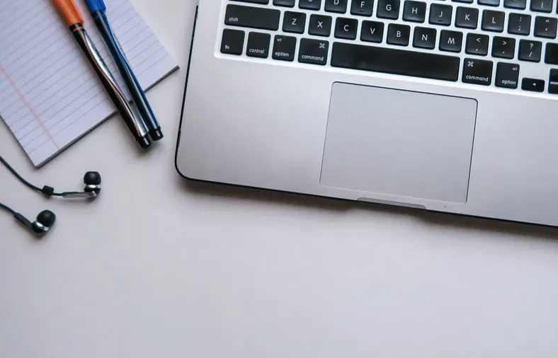 A laptop on a white desk beside a notepad, pens, and a pair of earphones neatly placed.