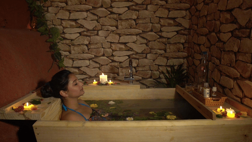 A woman relaxes in a wooden bathtub filled with water and flower petals, surrounded by candles, against a stone wall backdrop in a dimly lit, spa-like setting