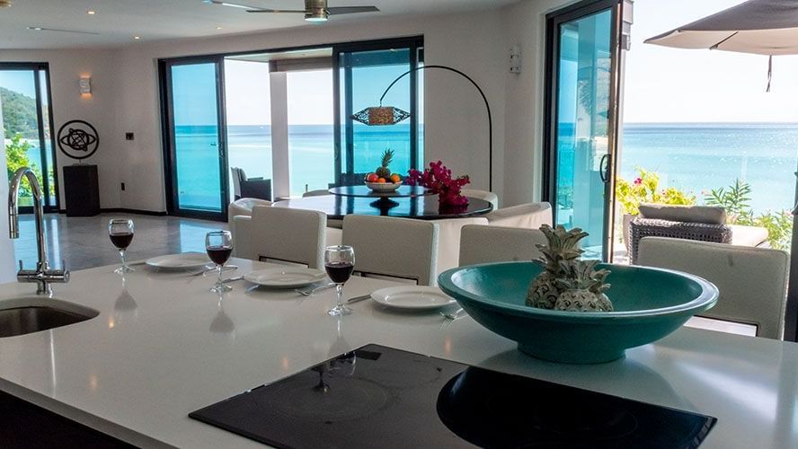 A modern kitchen island with place settings and wine glasses, looking into an open living and dining area with large glass doors that offer stunning views of the turquoise ocean.