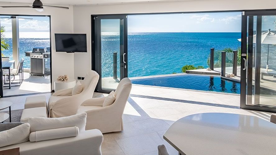 A bright, modern living room with white furniture opens up to an expansive ocean view through large glass doors, leading to an outdoor deck and turquoise water - Tamarind Hills Resort and Villas - Antigua