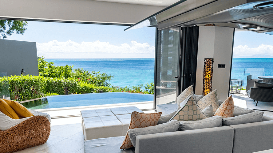 A spacious, modern living area with large windows opens up to a terrace with views of the ocean and lush greenery.