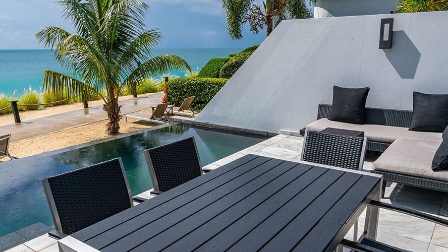 Private poolside patio with modern black furniture, lush greenery, and a stunning view of the ocean just beyond the villa garden.