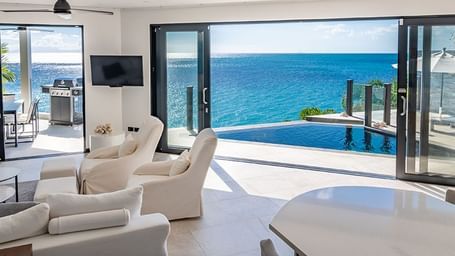 A bright, modern living room with white furniture opens up to an expansive ocean view through large glass doors, leading to an outdoor deck and turquoise water - Tamarind Hills Resort and Villas - Antigua