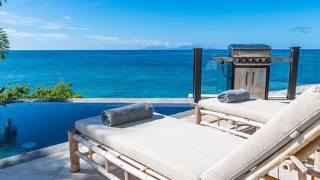 Rolled towels placed on sunloungers and an outdoor grill that are beside the deep blue infinity edge pools that seamlessly blends into the Caribbean Sea beyond Tamarind Hills Resort.