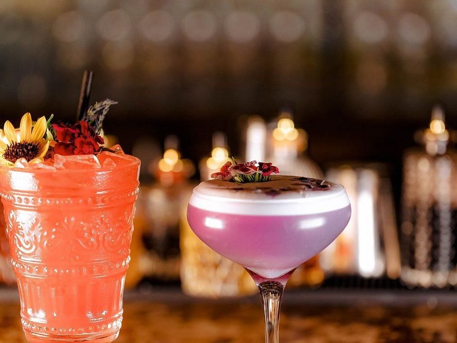 Glass filled with orange beverage topped with a flower accompanied by another glass filled with a lavender purple and pink beverage topped by flowers with a  blurred background.