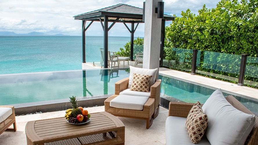 Luxurious patio with comfortable seating, a fruit bowl, and an infinity pool overlooking the turquoise ocean and a distant gazebo.