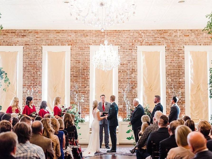 A grand wedding ceremony in a luxurious hall with tall windows, chandeliers, and an audience watching the couple exchange vows.- The Grand Hotel & Ballroom, McKinney