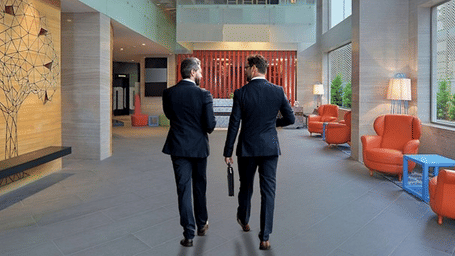 Creative corporate photography showing two businessmen in suits walking through the modern, spacious lobby of Radisson Bengaluru City Center Ulsoor Lake, featuring contemporary design elements and orange accent furniture, promoting the Business Class Room 