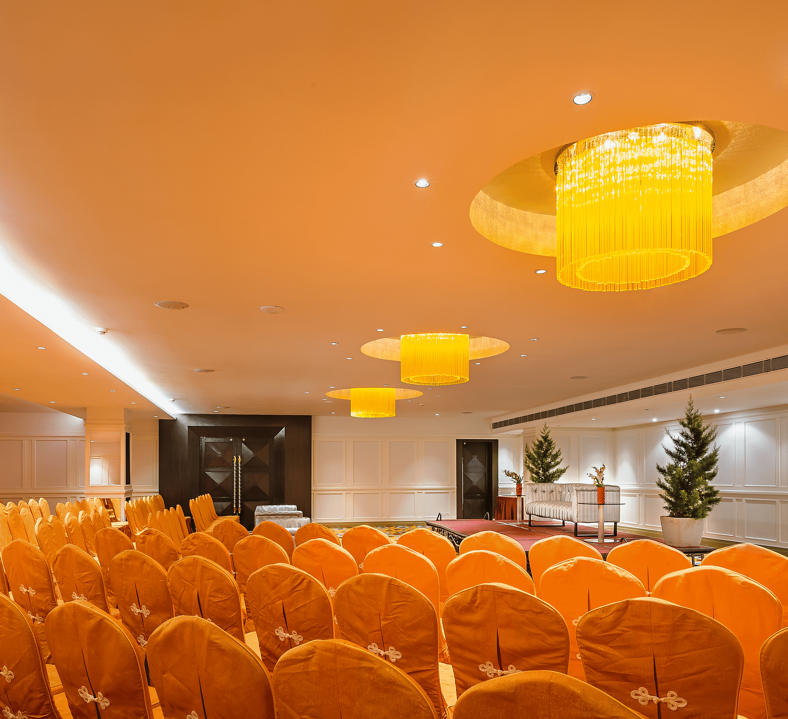 Brightly lit grand ballroom with yellow lighting, white walls, orange seating and a stage with a Christmas tree and red carpet - TGI Grand Fortuna, Hosur