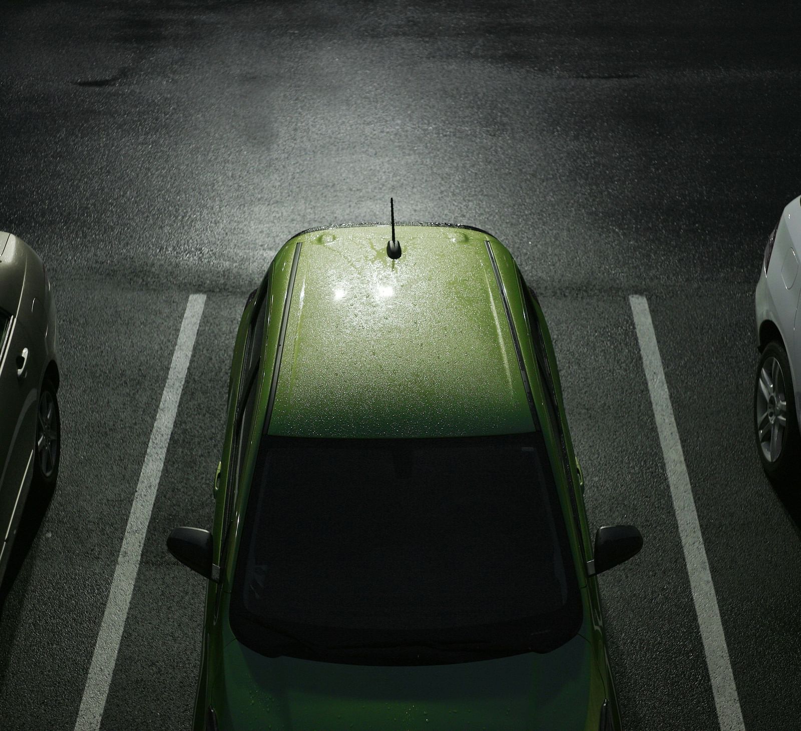 An overhead view of a parking lot at night with a lime green car between two white cars under streetlights