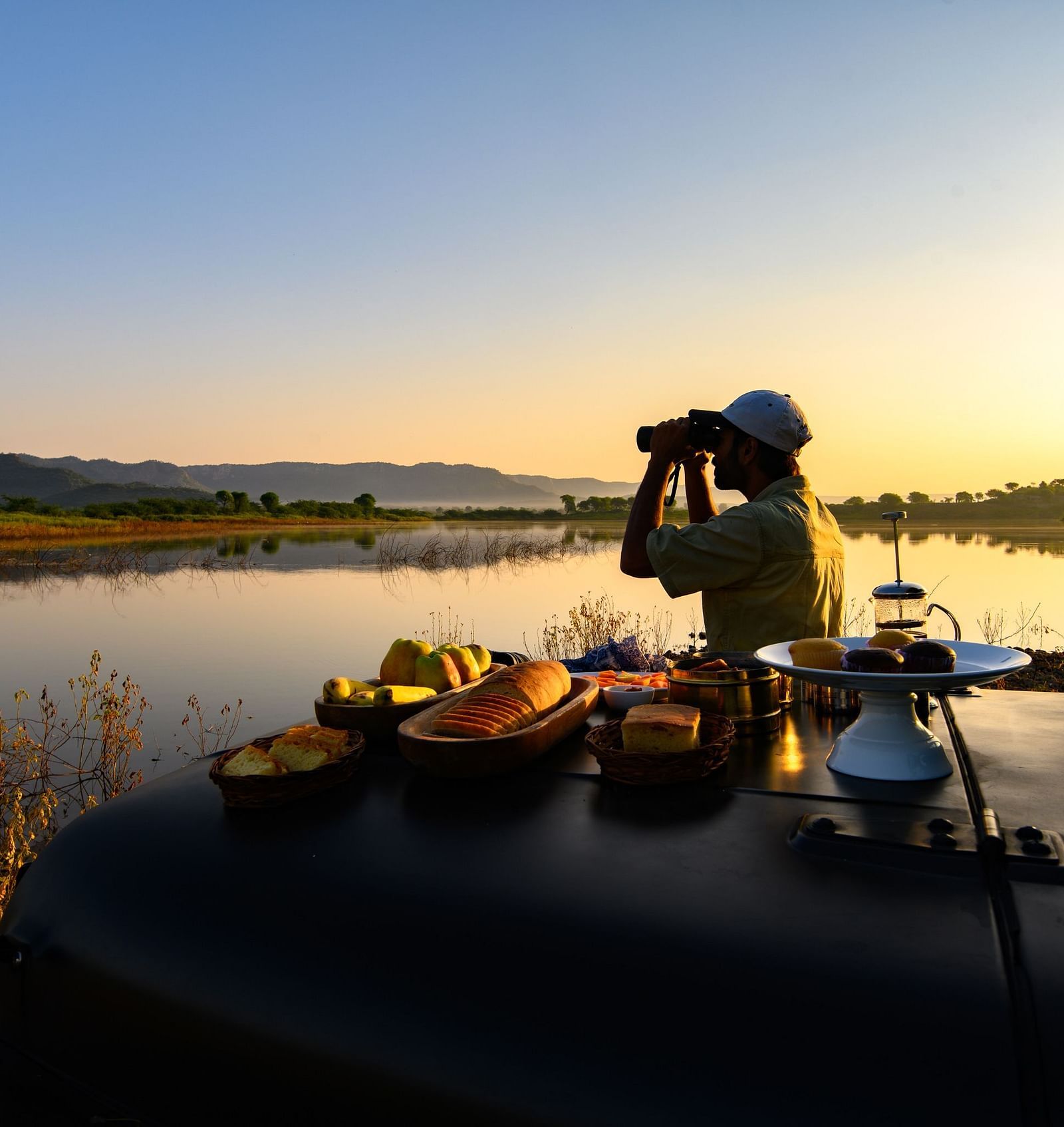 An image of a man standing near a lake with his jeep parked over looking the mountain - An image of a jeep parked amidst the rocky mountains - Utsav Camp Sariska