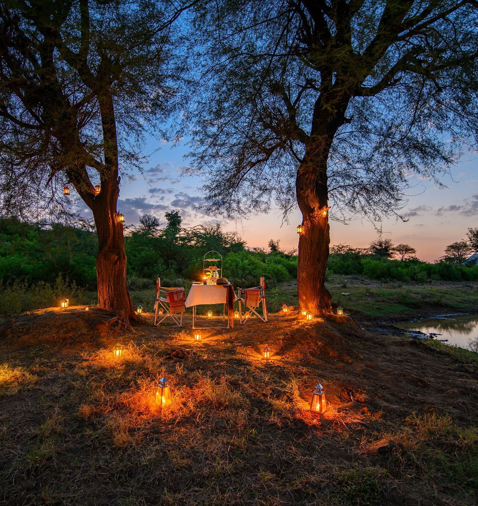 An image of a dining set up near a lake with decorative lighting and trees and greenery - Utsav Camp Sariska