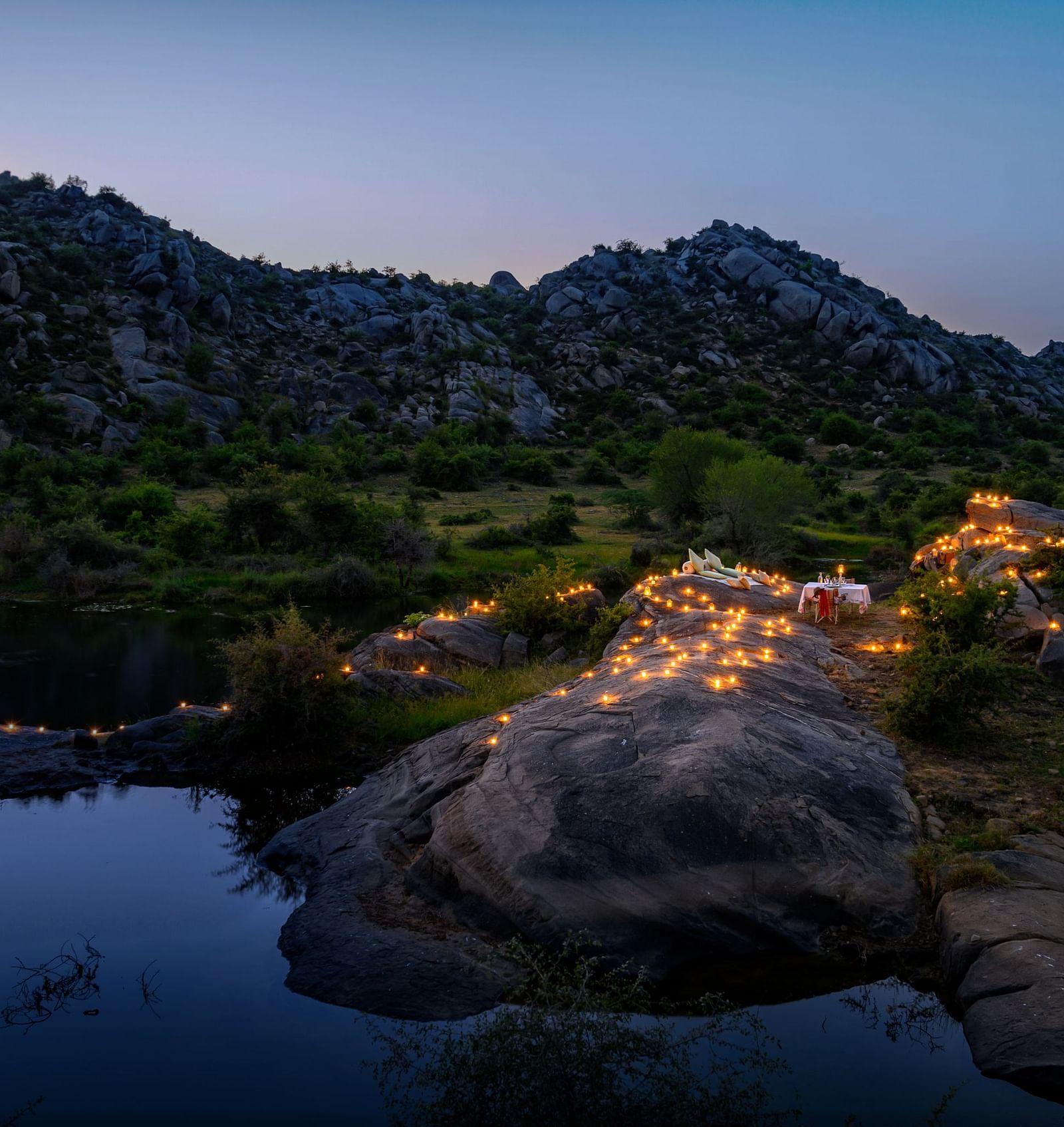 An image of an outdoor dining arrangement near a lake over viewing the mountains with bright lighting and greenery around - Utsav Camp Sariska